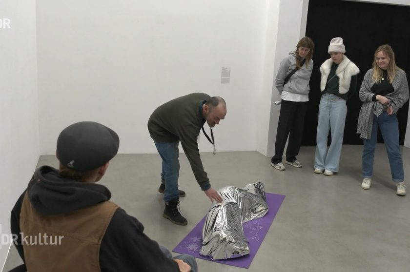 In einer Galerie steht eine Person, die sich &uuml;ber eine mit einer silbernen Rettungsdecke bedeckte Figur auf einem lila Teppich beugt, w&auml;hrend vier weitere Personen zuschauen.