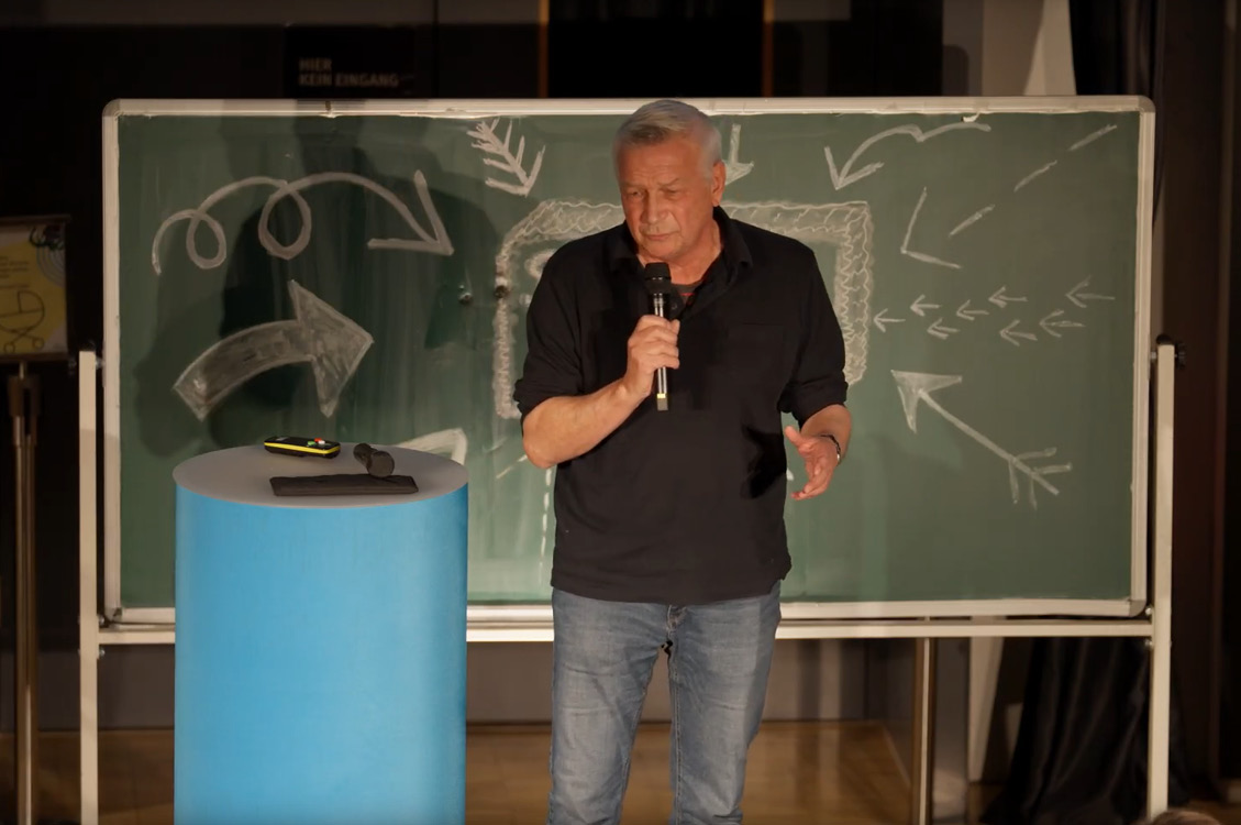 Mann steht vor einer Tafel mit Pfeilzeichnungen und hält ein Mikrofon, neben ihm ein runder blauer Stehtisch mit Gegenständen