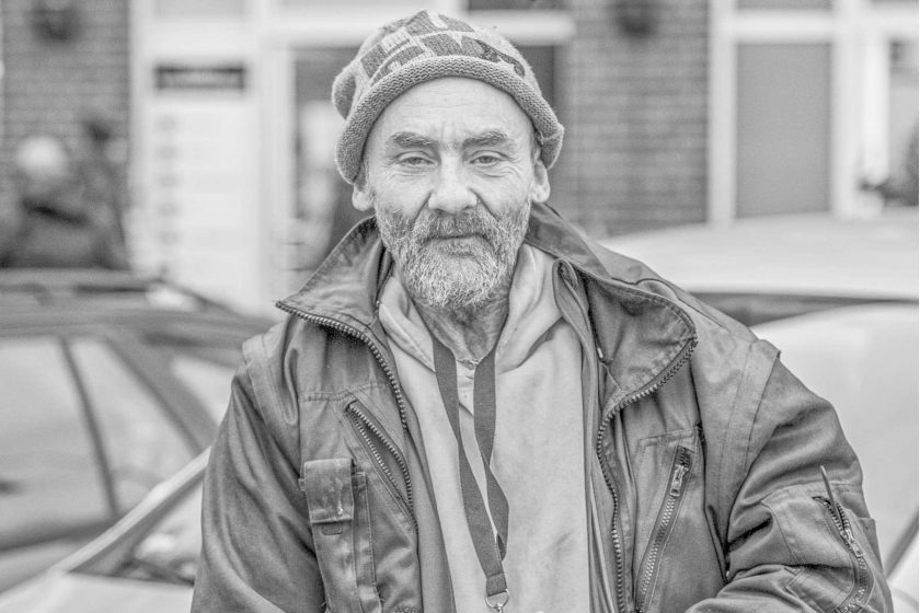 Mann mit kariertem M&uuml;tze, Jacke und Pullover steht vor einem geparkten Auto in einer urbanen Umgebung