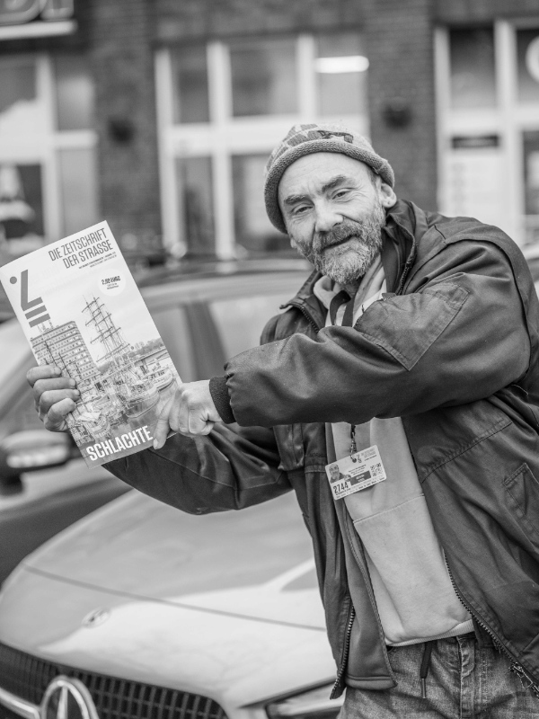 Mann mit Mütze und Jacke hält eine Zeitung mit dem Titel 'DIE ZEITSCHRIFT DER STRASSE' vor einem geparkten Auto hoch